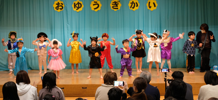 つくし保育所のお遊戯会の模様の模様。園児12名が個性豊かなきらびやかな衣装をまとい踊っている姿
